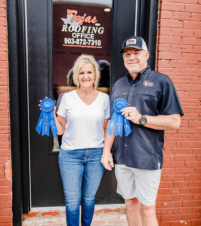 Owners, David and Phyliss Jones, standing in front of the Tejas Roofing & Gutters office building holding '2021 Corsicana Best of the Best' blue ribbon awards